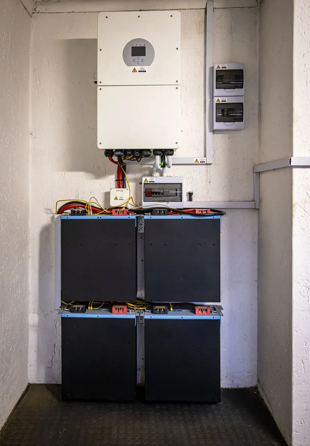 A wall-mounted inverter above a stack of four battery modules, with isolator switches and cabling