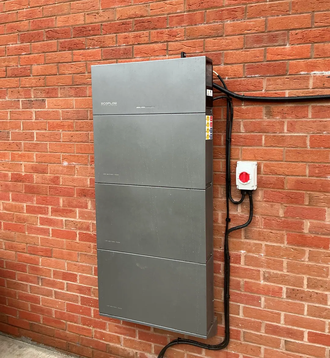 An EcoFlow hybrid inverter above a stack of three battery packs on an outside brick wall, a red isolator switch beside them