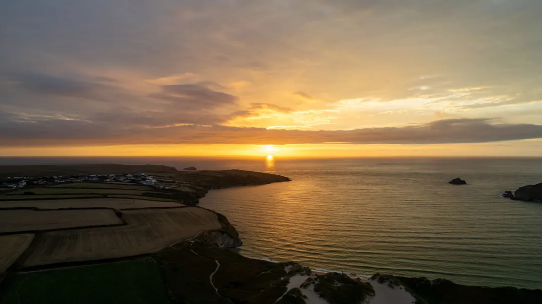 Aerial view of fields and a village on a headland above the sea on the Cornish coast, with the sun setting over the water