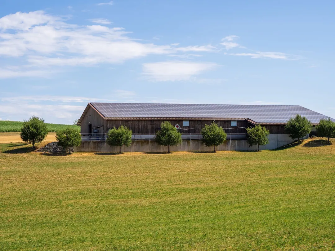 A long timber barn with a solar array across its roof, set among green fields