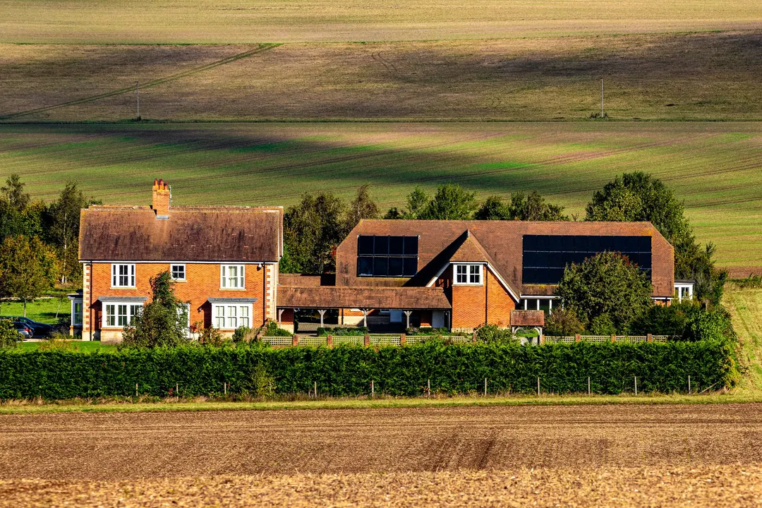 A red-brick country house with black solar panels on its roof, surrounded by fields