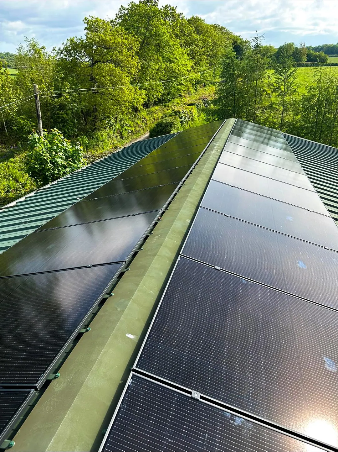 Black solar panels running the length of a green metal barn roof, both slopes, with trees and fields beyond