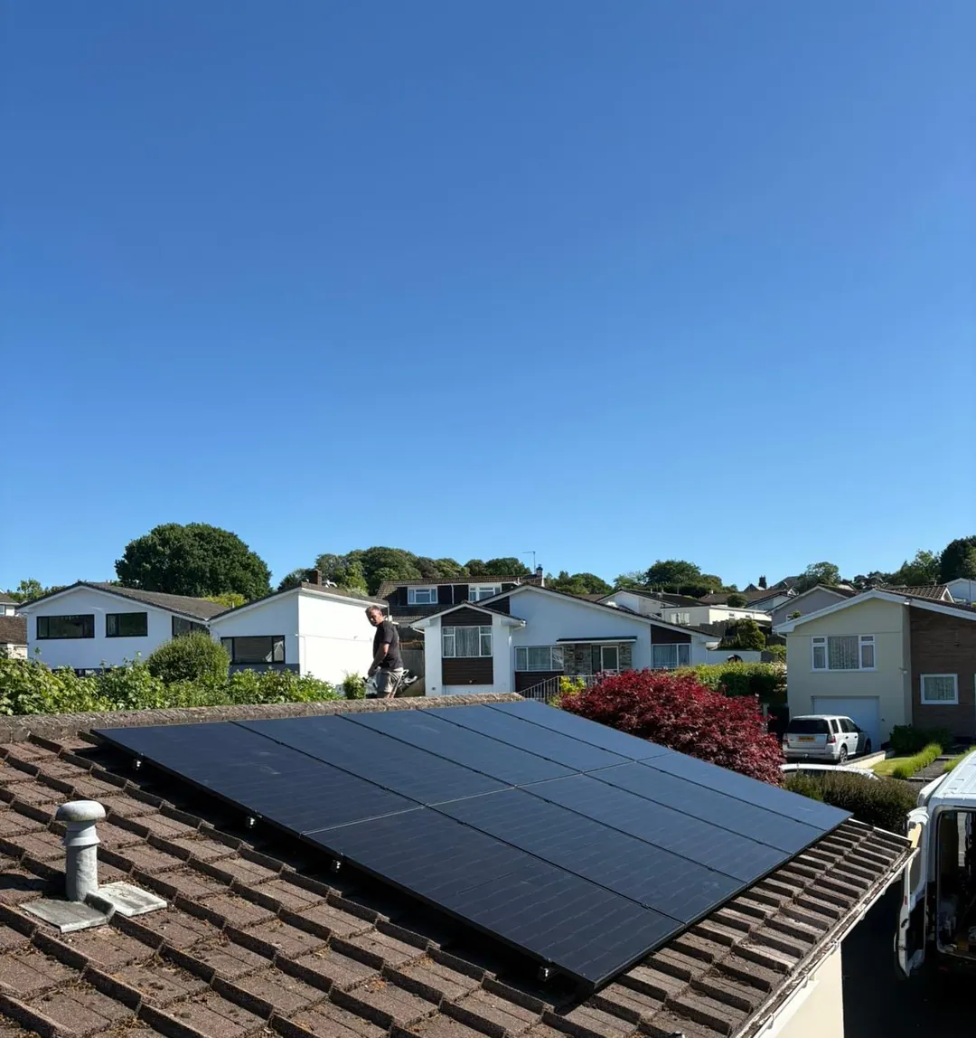 A black solar array on a bungalow’s tiled roof, with an installer on the roof behind and houses beyond