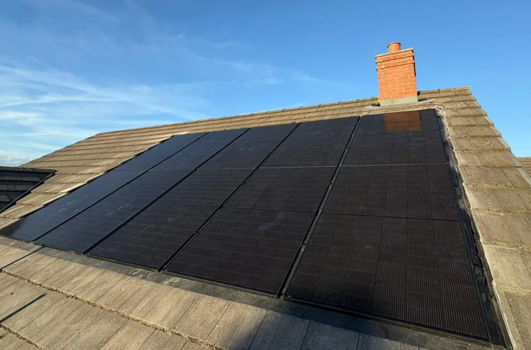 An all-black solar array on a concrete-tiled roof below a red-brick chimney, under a blue sky