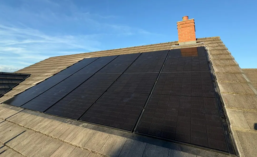 Black solar panels across a concrete-tiled roof slope below a red-brick chimney, under a blue sky