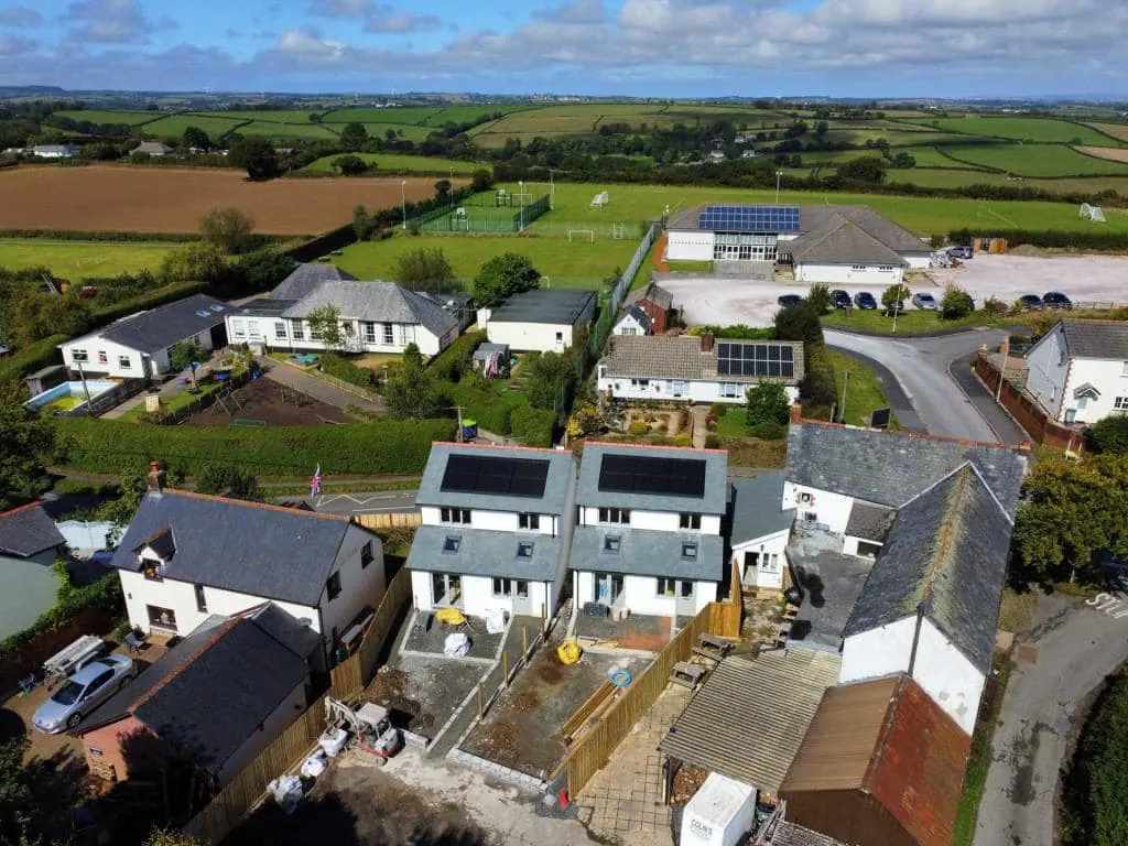 A drone view over a village: two new-build houses with black solar panels on their slate roofs, fields and playing fields beyond