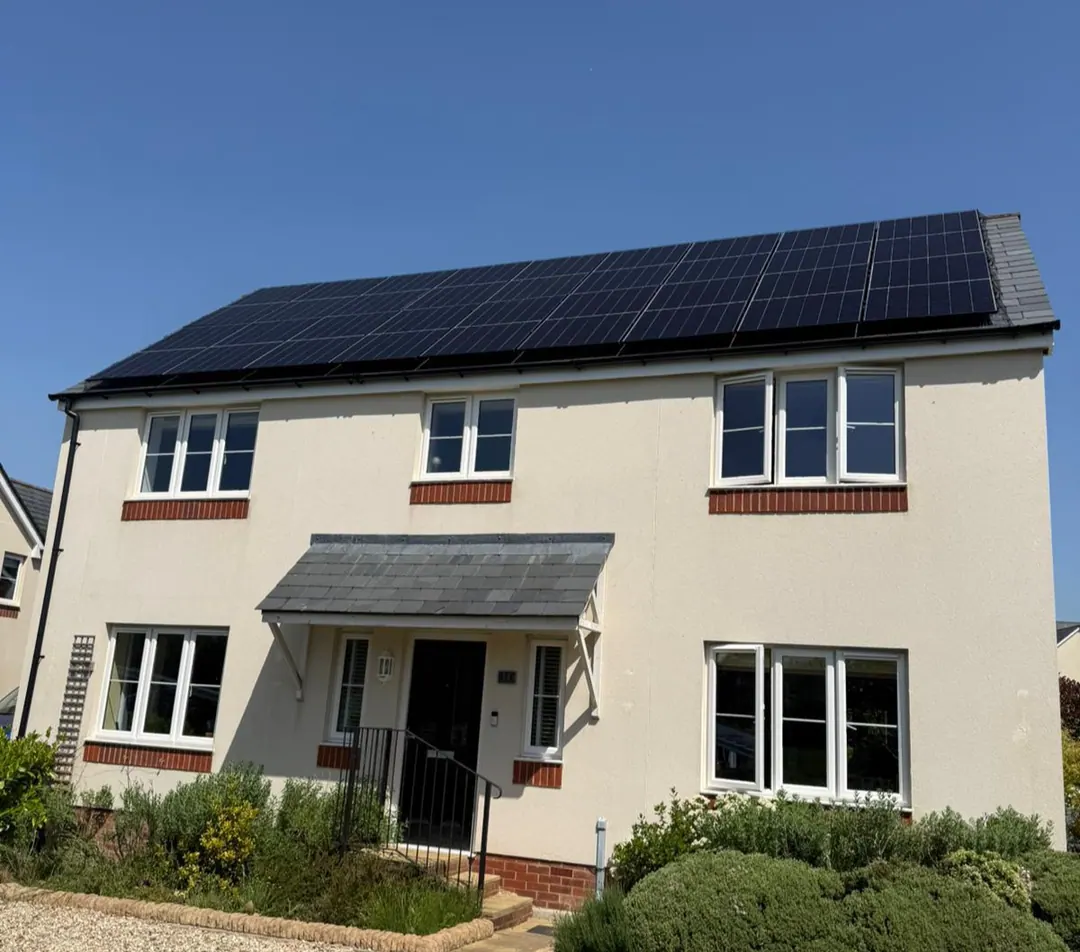 A cream-rendered detached house with a row of black solar panels across its roof, under a clear blue sky