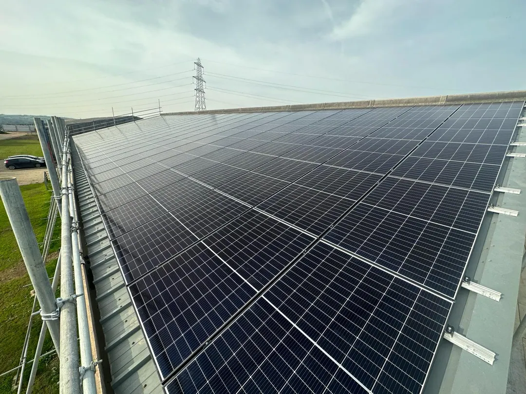 Rows of solar panels across a long sheeted roof, with scaffolding along its edge and an electricity pylon beyond