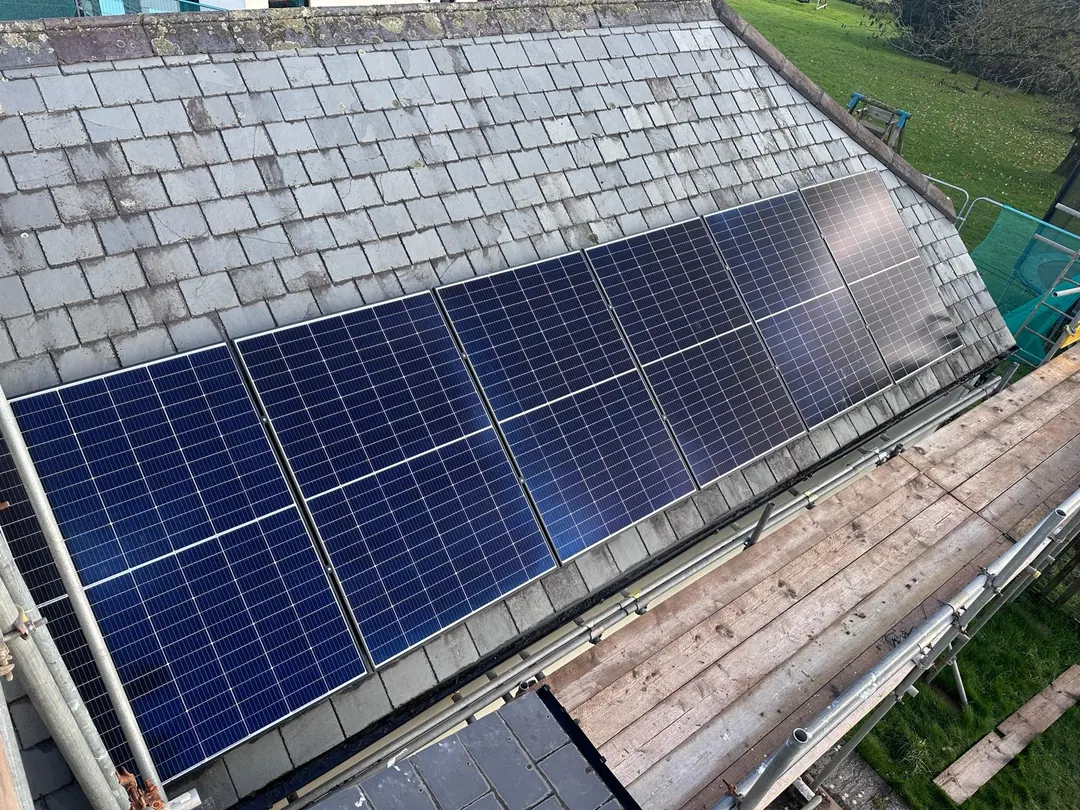 A single row of blue solar panels on an old slate roof, seen from above, with scaffold boards below and a lawn beyond