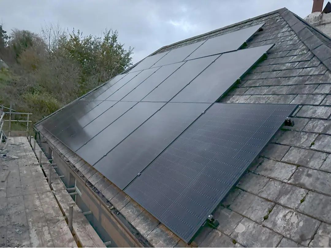 Black solar panels fixed above grey slates in two staggered rows, with scaffolding along the eaves under a cloudy sky