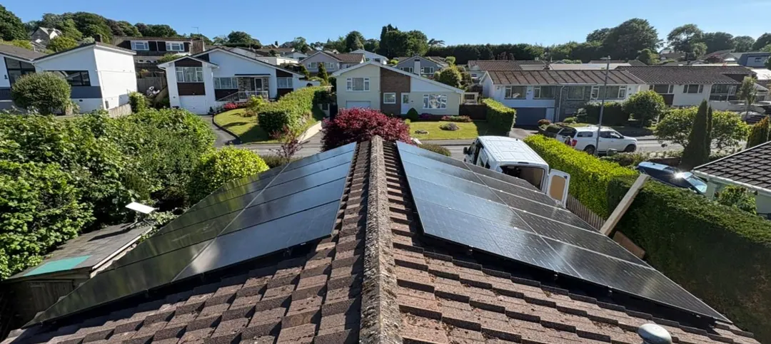 Black solar panels on both slopes of a tiled roof, seen from above the ridge, with a street of houses and gardens beyond